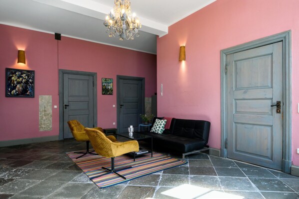 A hallway with a pink wall, a black sofa, a yellow chair, a chandelier, and two doors. - Steffenshagen Vicarage Retreat (Steffenshagen)