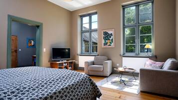 Ein Zimmer mit einem Bett, einem Fernseher, einem Sofa und einem Sessel. Große Fenster und eine Tür sind vorhanden.