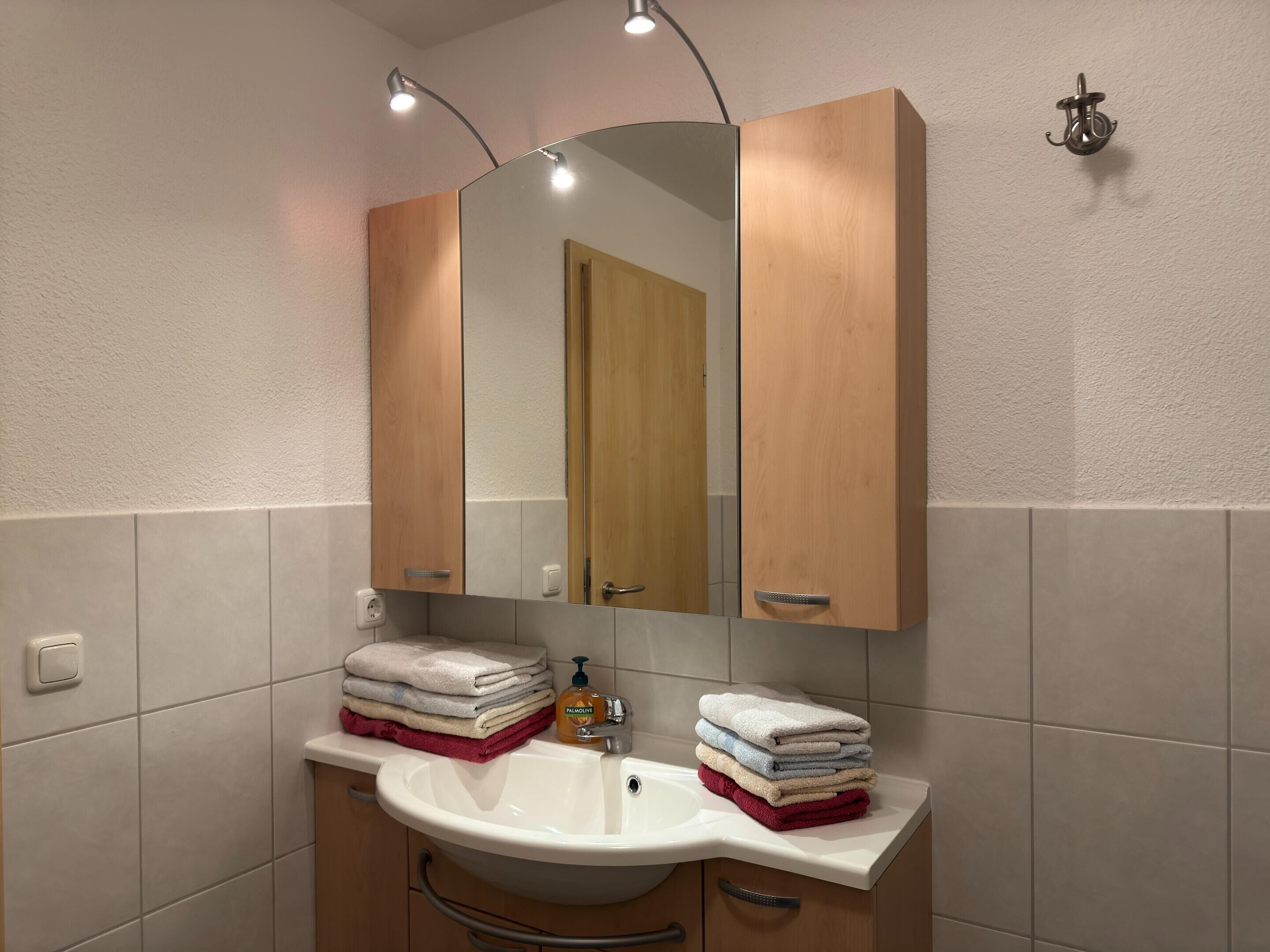 Un baño con un lavamanos blanco, armarios de madera y un espejo encima. Hay toallas apiladas en el lavamanos y un dispensador de jabón en la encimera.