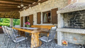 Patio esterno con pavimento in pietra, tavolo in legno, sedie in metallo e camino in pietra.