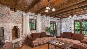 Una stanza accogliente con pareti in pietra, travi in legno, un camino in pietra, un divano, un tavolino da caffè e ampie finestre.