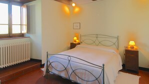 Ein Schlafzimmer mit einem Metallbettgestell, zwei Nachttischlampen, einem Heizkörper und einem Fenster mit Jalousien.