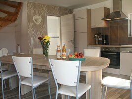 A modern kitchen with a wooden dining table, white chairs, a vase with flowers, and a fruit bowl.
