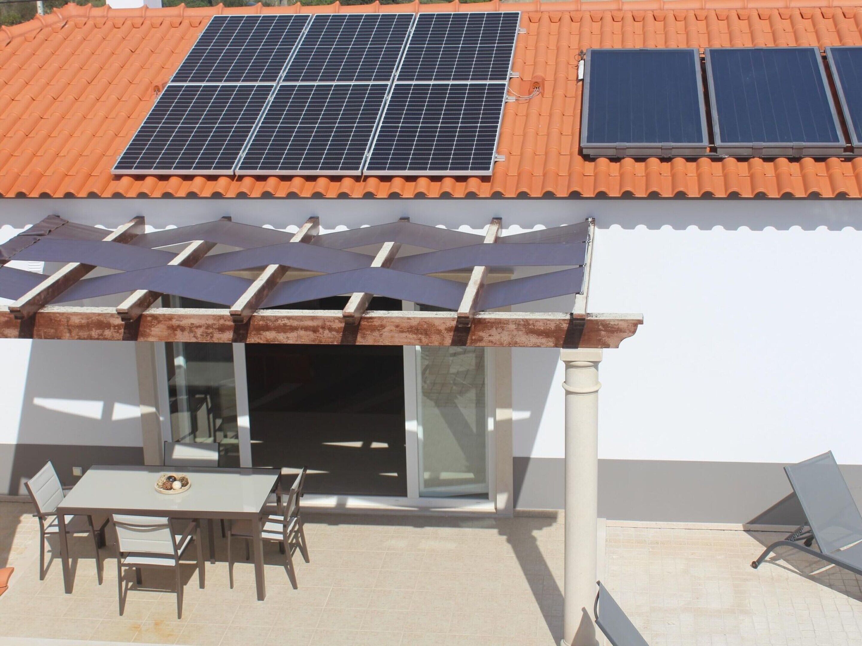 Un edificio con techo de tejas, paneles solares, un porche cubierto con mesa y sillas, y un acceso de vidrio a un espacio interior.
