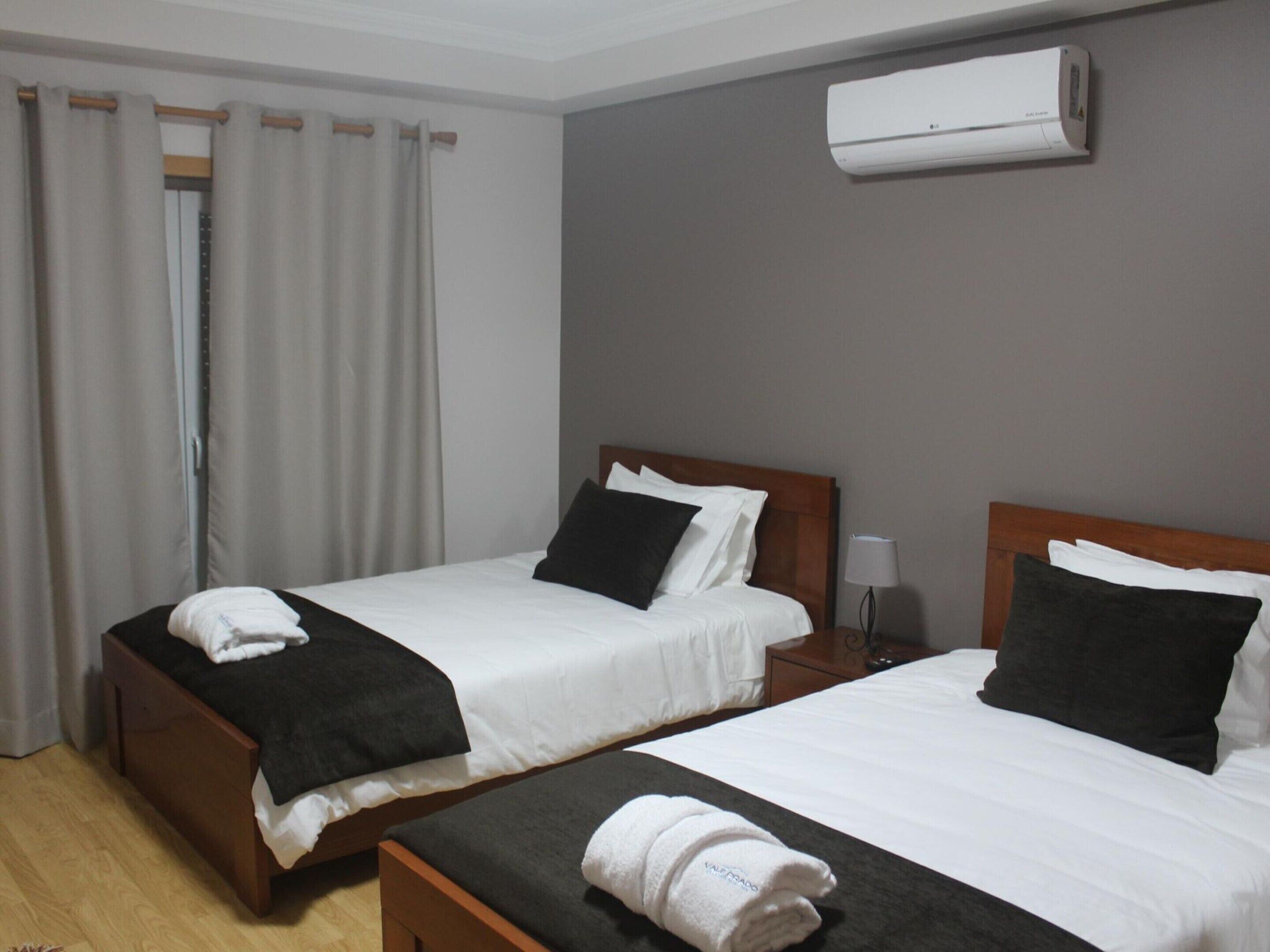 Habitación de hotel con dos camas, cabecera de madera, mesita de noche, lámpara y unidad de aire acondicionado.