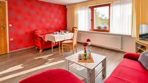 A room with a red sofa, a dining table with chairs, a small coffee table with a remote control, and a window with curtains. - Apartment in Klutz Near Baltic Sea Beach (Klütz)