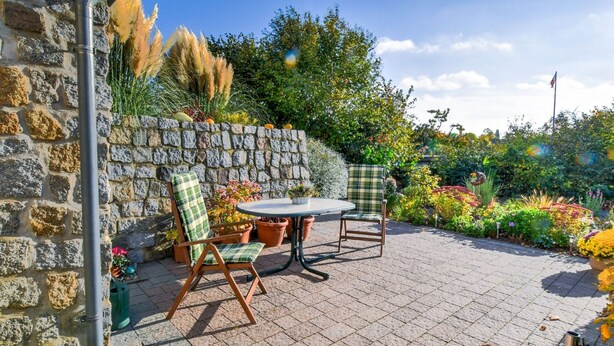 A patio with a stone wall, two chairs, a table, and a variety of plants and flowers. - Apartment in Klutz Near Baltic Sea Beach (Klütz)