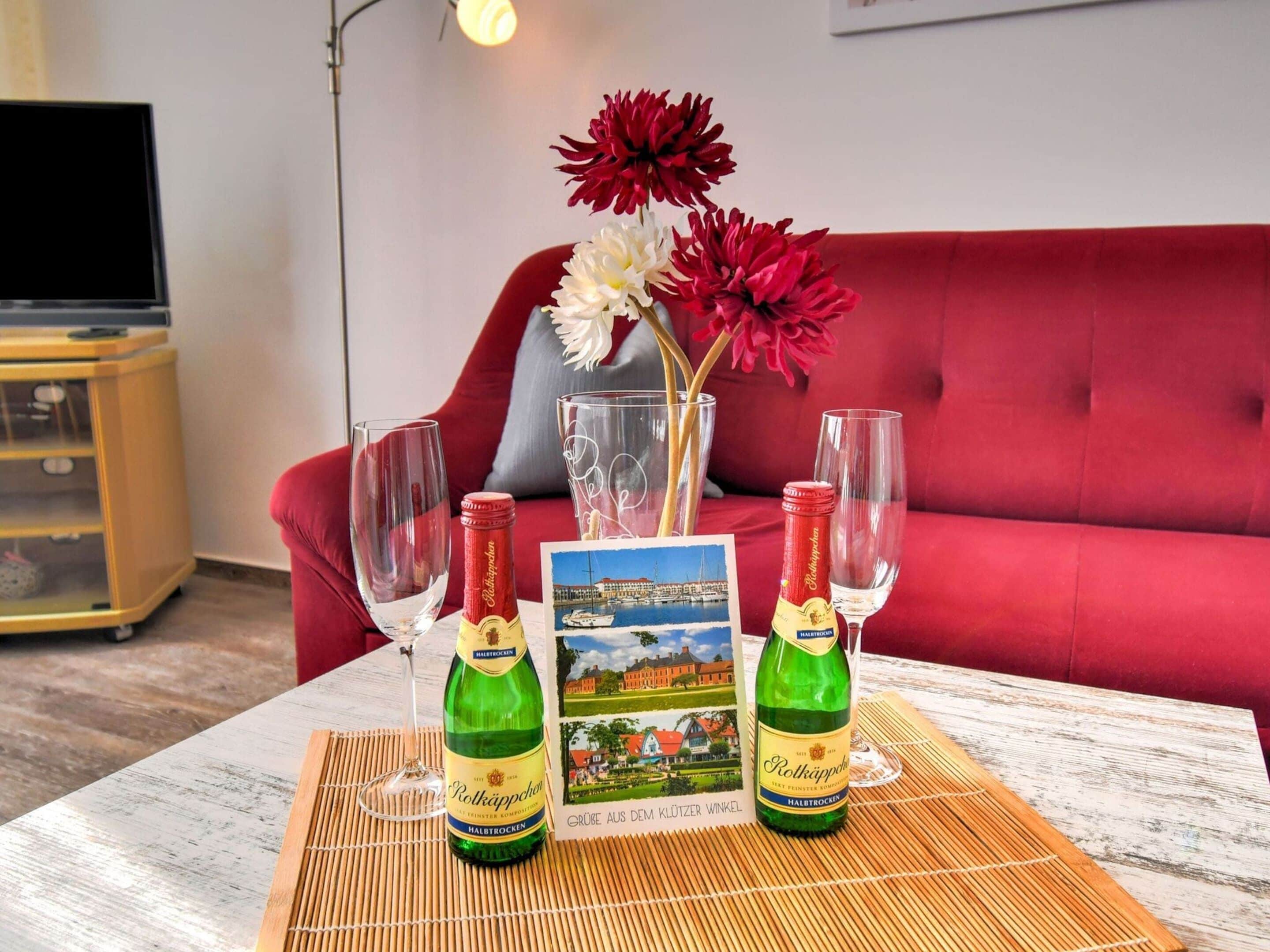 A table with two bottles of Rottkappfen beer, two wine glasses, a vase with red and white flowers, and a postcard of a scenic view.
