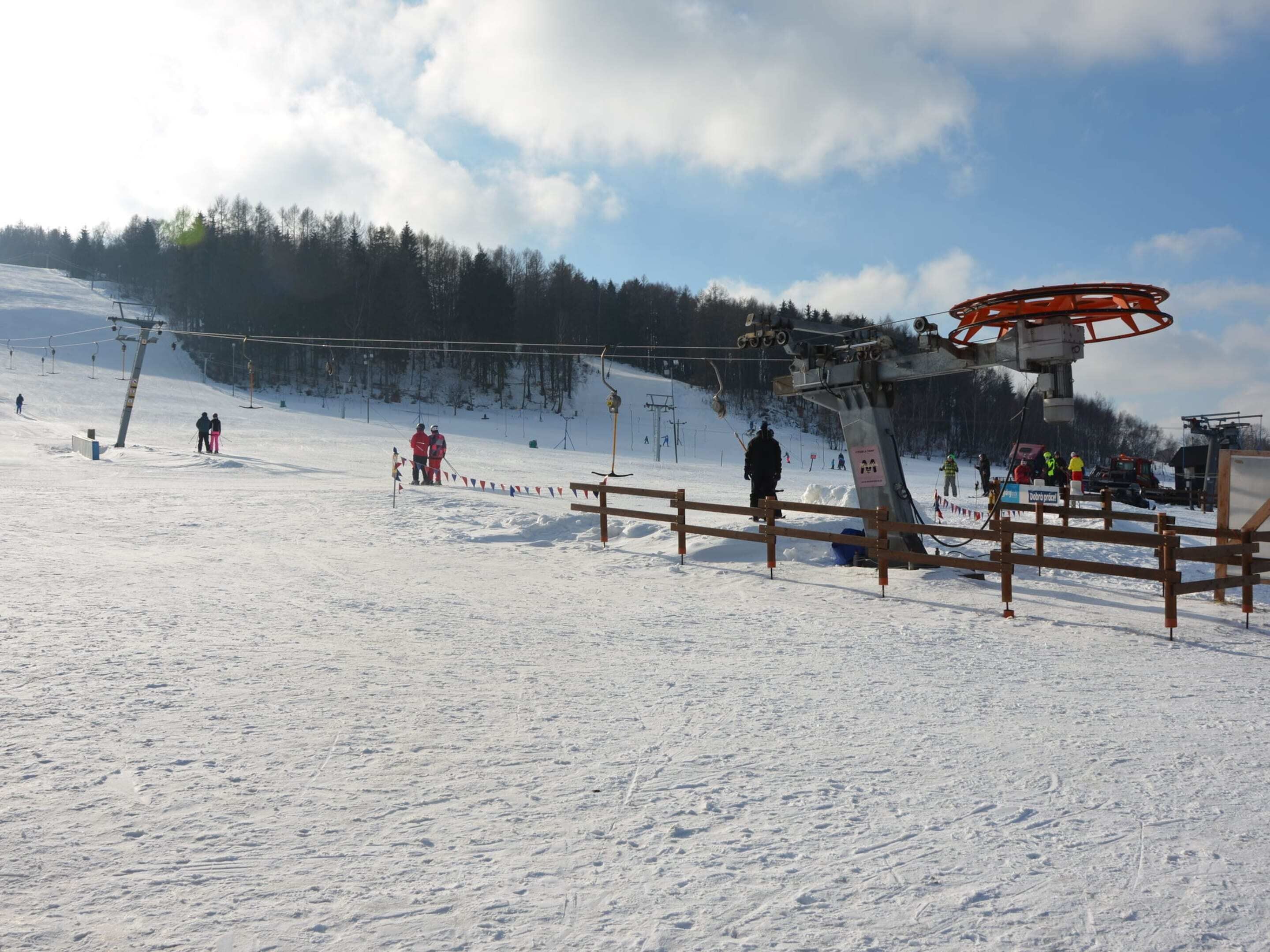 A ski lift with a wooden fence in the foreground, a snowy slope, and a forested hill in the background.