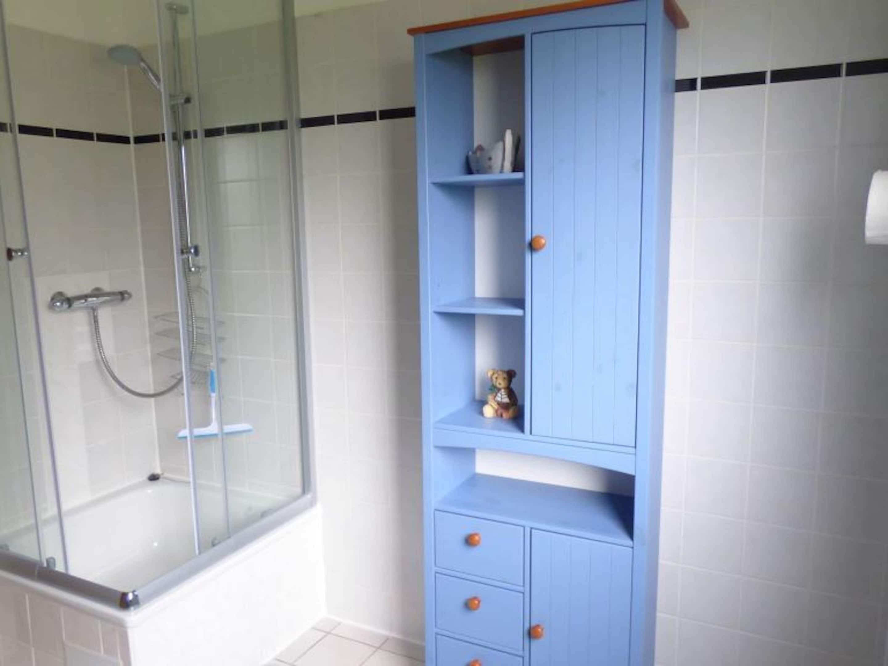 A bathroom with a glass-enclosed shower, white tiled walls, and a blue cabinet with drawers.