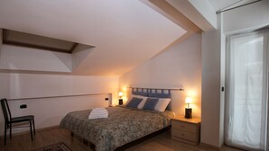 A bedroom with a sloped ceiling, a bed with a headboard, bedside tables with lamps, a chair, a rug, and a window with curtains. - Apartment in Predazzo Near Dolomiti Superski (Predazzo)