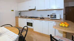 A compact kitchen with white cabinets, a microwave, a gas stove, and a small sink. There's a dining table with chairs and a bowl of fruit on the counter. - Apartment in Predazzo Near Dolomiti Superski (Predazzo)