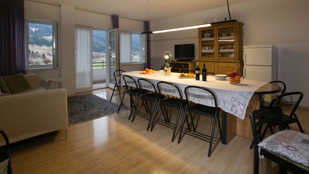 A modern living room with a sofa, a dining table with chairs, a kitchen area with a TV and cabinets, and a view of the outdoors through large windows. - Apartment in Predazzo Near Dolomiti Superski (Predazzo)