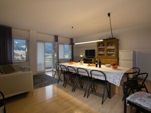 Un saló modern amb un sofà, una taula de dinar amb cadires, una zona de cuina amb televisió i armaris, i vista a l'exterior a través de grans finestres.
