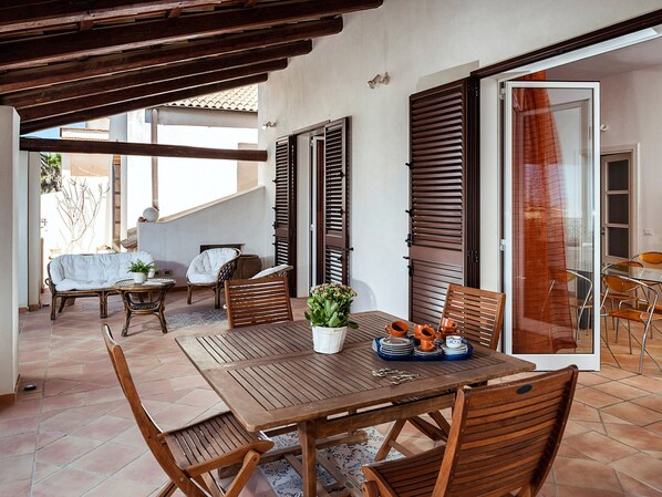 Un patio exterior con una mesa y sillas de madera, una planta en maceta y un ventanal de vidrio que da a un espacio interior.