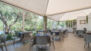 A covered outdoor dining area with tables and chairs, large windows, and a view of greenery.