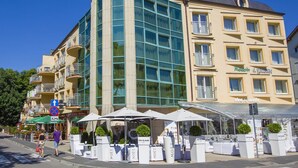 Un hôtel doté d’une façade moderne en verre, d’un espace extérieur aménagé avec des parasols et d’une enseigne indiquant « Viva la moda ».