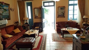 A cozy living room with a brown sofa, a matching ottoman, a coffee table, and a patterned rug. There is a door leading outside, a window with curtains, and a wall-mounted lamp. - Polyxenia Hotel (Nafplio)