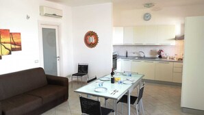 Uma sala moderna com um sofá marrom, uma mesa de jantar com cadeiras, área de cozinha com armários brancos, um relógio e um espelho decorativo.