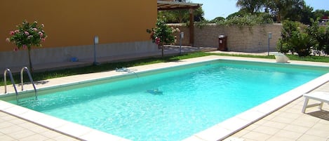 A rectangular swimming pool with a tiled deck, a pergola with a roof, and a stone wall in the background.