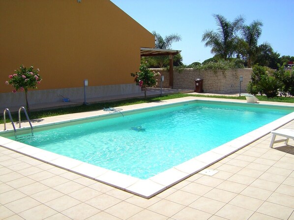 Ein rechteckiger Pool mit Fliesen umgeben, eine Pergola mit Dach und eine Steinmauer im Hintergrund.