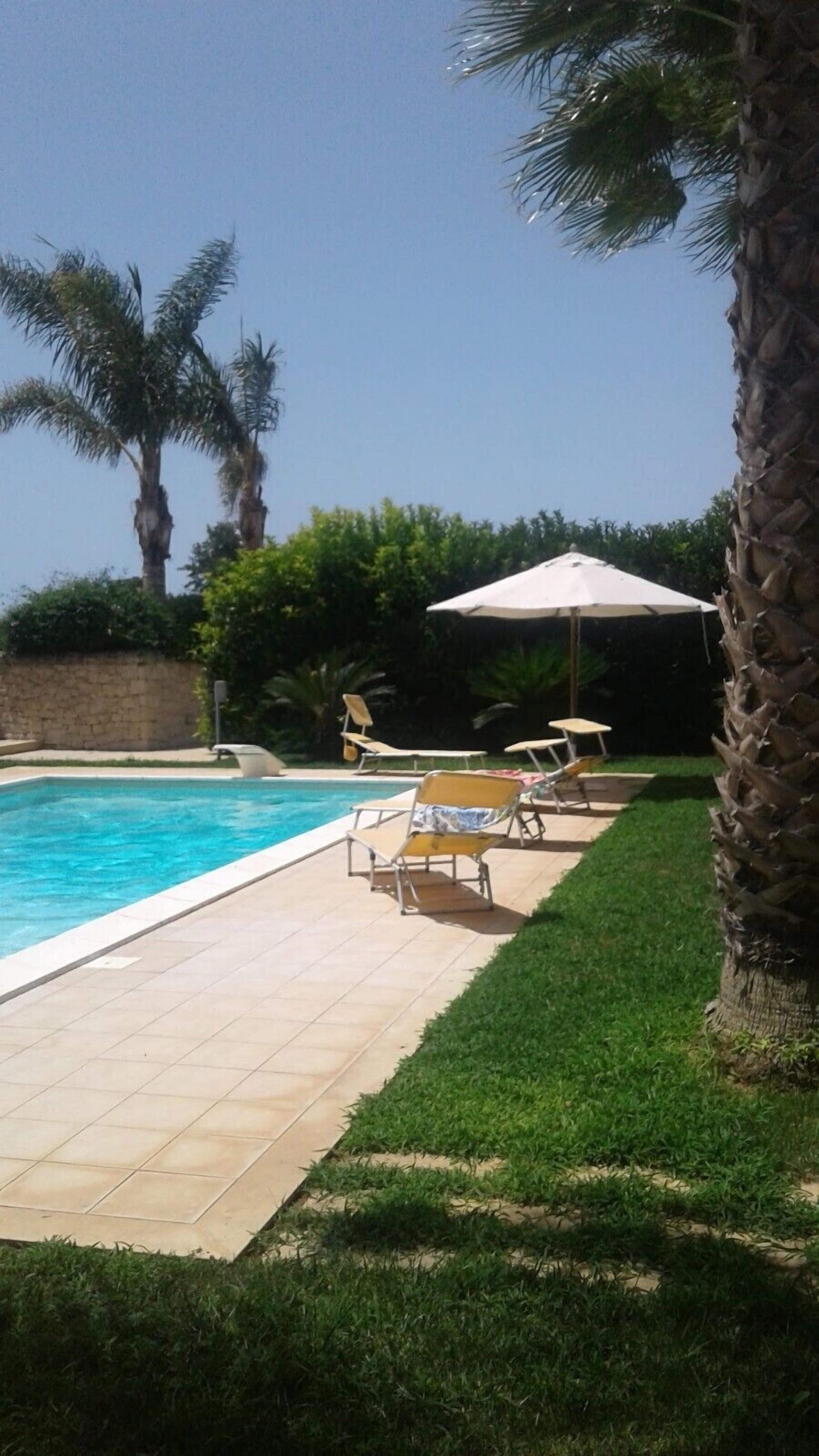 Une piscine dont le fond est recouvert de carrelage, entourée de palmiers et bordée d’une pelouse où se trouvent des chaises longues et un parasol.