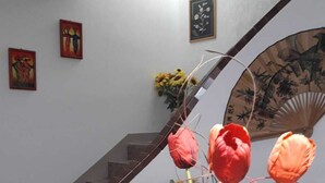 Een eetgedeelte met een rieten stoel, een tafel met een wit tafelkleed en een vaas met bloemen. Op de achtergrond is een trap met houten leuning zichtbaar.