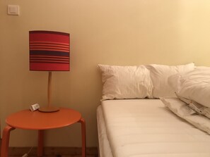 Una cama con ropa blanca, una lámpara a rayas rojas y negras sobre una mesita naranja, y una pared blanca.