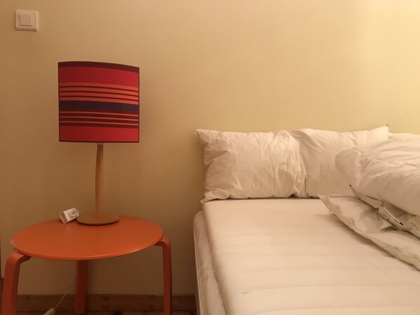 Un lit avec une literie blanche, une lampe à rayures rouges et noires posée sur une petite table orange, et un mur blanc.