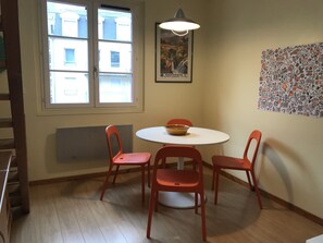 A dining area with a round table, four red chairs, a bowl on the table, a window with a view of buildings, and a poster on the wall. - Coeur de Cauterets, cosy T2 + mezzanine pr 4 Pers (Cauterets)