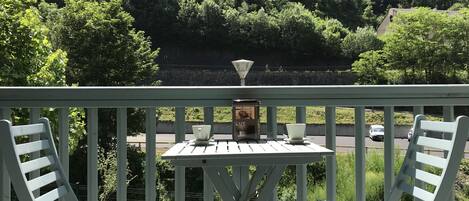 A balcony with two white chairs and a table, overlooking a road and dense greenery.