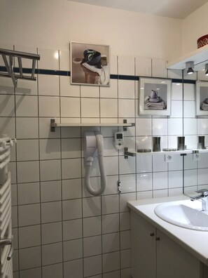 Kylpyhuoneessa on valkoinen pesuallas, pyyhekoukku, letkulla varustettu suihku ja seinällä kehyksiin kiinnitetyt valokuvat.