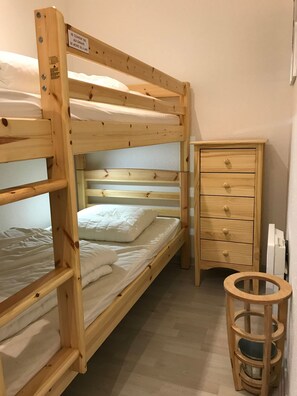 Una cama litera con estructura de madera, un pequeño aparador de madera con cajones y un taburete redondo de madera.