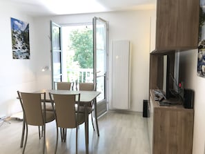 Una habitación con una mesa de comedor y sillas, un televisor sobre un mueble de madera, y un ventanal con vistas a zonas verdes.