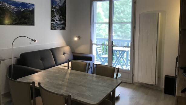 A dining area with a table and chairs, a sofa, a pendant light, and a window with a view of greenery. - T2 +alcove, récent 4/5 pers. Parking. Balcon. Wifi (Cauterets)