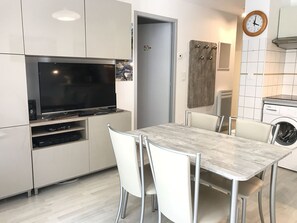 Moderni keittiö, jossa on litteänäyttöinen televisio, integroidut kodinkoneet sekä ruokailutila pöytineen ja tuoleineen.