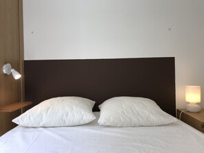 A neatly made bed with two pillows, a wooden headboard, a bedside lamp, and a wall-mounted light fixture.