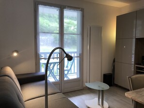 A compact living space with a sofa, a small table, a cabinet, and a window with a view of greenery. - T2 +alcove, récent 4/5 pers. Parking. Balcon. Wifi (Cauterets)