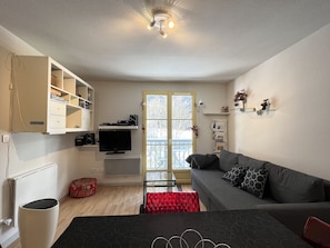 A living room with a grey sofa, a TV, a bookshelf, and a window with a view of trees.