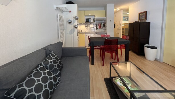 A modern living room with a grey sofa, a dining table with red chairs, a kitchen in the background, and a small trash bin. - T2 alcove confortable pour 4 à 5p. Balcon. Parking (Cauterets)