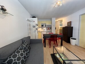 Un espacio habitable compacto con un sofá gris, una mesa de comedor con sillas rojas, una zona de cocina y un pequeño cubo de basura.
