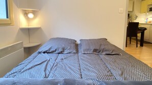A small, single-bed room with a bedside table, a chair, and a view into a kitchen area. - T2 alcove confortable pour 4 à 5p. Balcon. Parking (Cauterets)