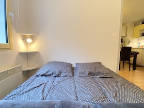 A small, single-bed room with a polka dot bedspread, a bedside lamp, and a view into a kitchen area.