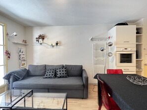 Una sala moderna con un sofá gris, una mesa de centro de vidrio y una mesa de comedor con una silla roja. Hay un armario blanco con horno incorporado y una repisa de vidrio con objetos decorativos.
