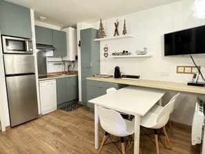 Moderni keittiö, jossa on sisäänrakennettu mikroaaltouuni, uuni ja astianpesukone. Keittiössä on puinen ruokapöytä, valkoiset tuolit ja pöydän yläpuolella pieni hylly.