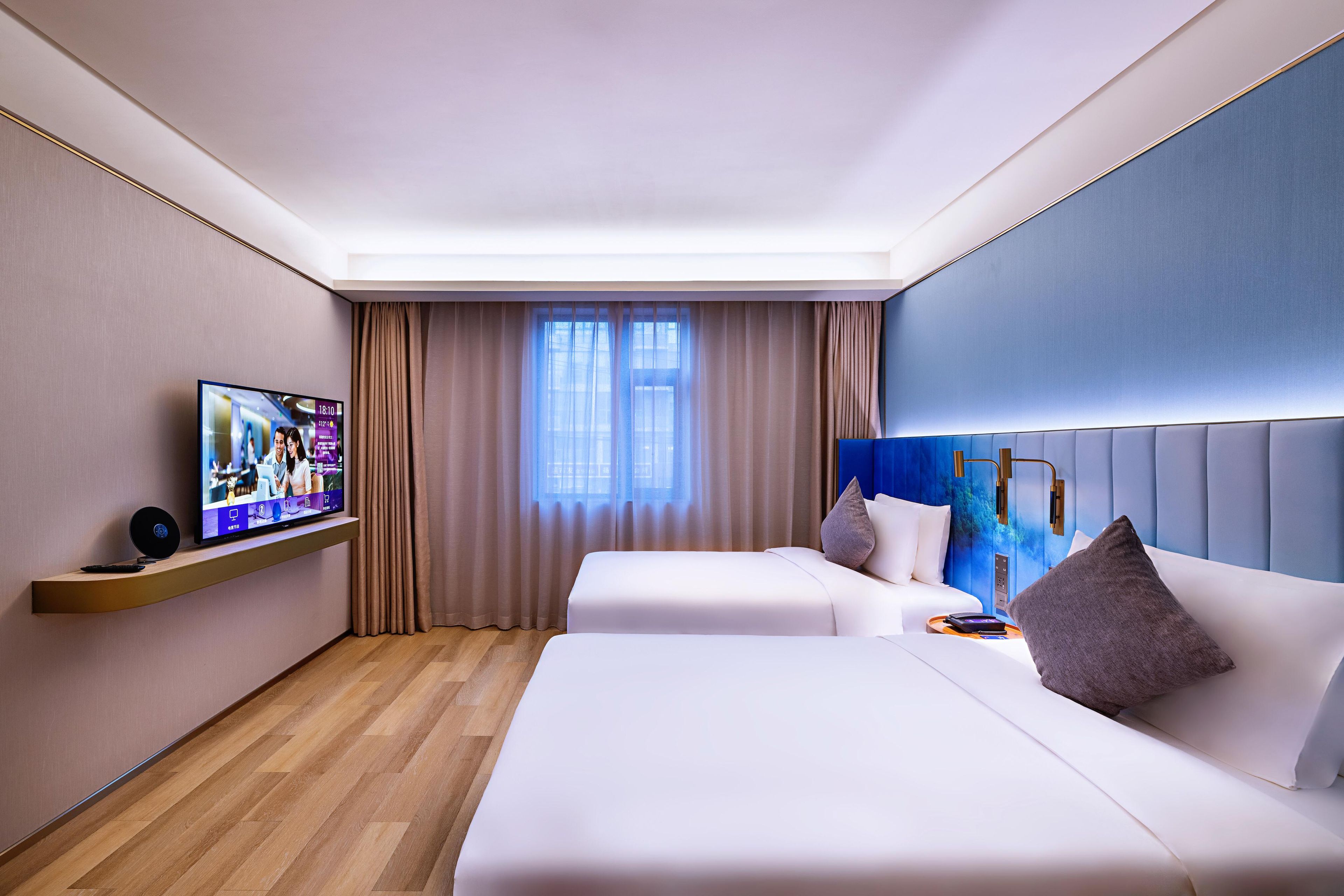 A hotel room with a television on a shelf, two beds, a headboard, and a window with curtains.