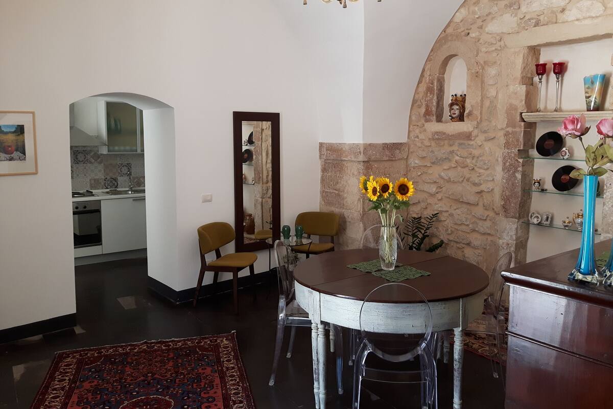 A dining area with a round table, chairs, and a vase with sunflowers. There is a stone wall and a shelf with decorative items.