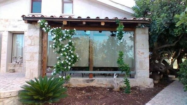 Un edificio in pietra con tetto in tegole, un'ampia finestra adornata da una vite con fiori bianchi, e un piccolo giardino con una pianta e una panchina.