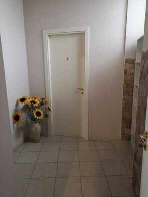 Um corredor com uma porta branca, piso de azulejos e um vaso com girassóis à esquerda.