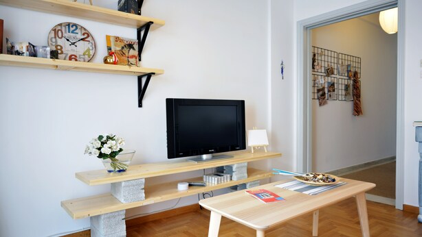 Una sala de estar con un televisor de pantalla plana, estantes de madera, un banco de hormigón, una mesita y un jarrón de flores.
